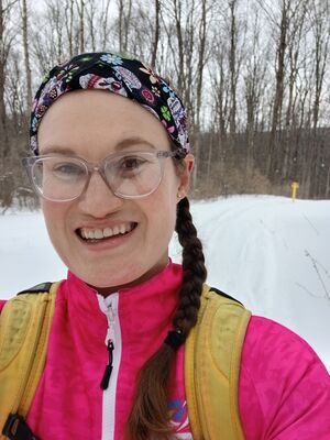 An image of Maryellen Tighe in the woods cross country skiing. She is wearing a neon pink jacket with a neon yellow backpack and a bandana with a dia de los muertos skull design.